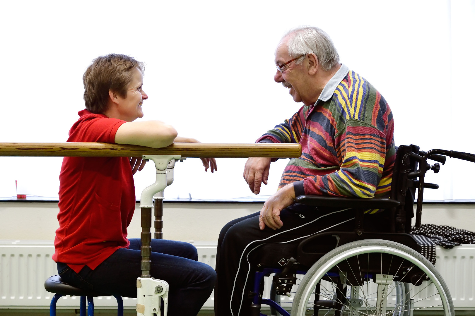 Fysiotherapeut zit op kruk in gesprek met patiënt in rolstoel
