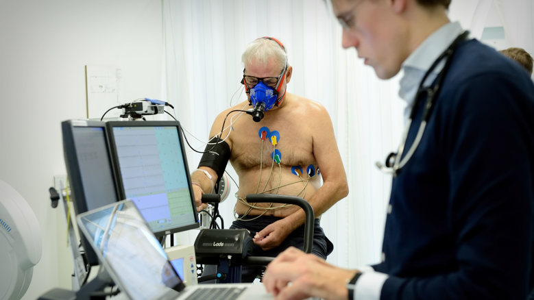 Een patient zit op een fietsergometer voor de inspanningstest terwijl de arts de uitslagen van de meetinstrumenten in de gaten houdt