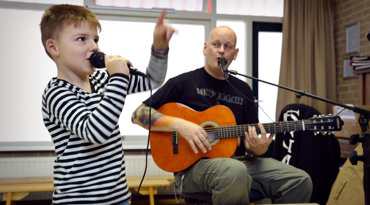 Leerling zingt in een microfoon en leeraar begeleid hem met de gitaar